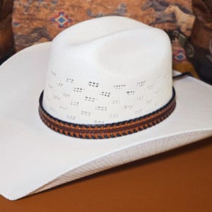 May include: A white cowboy hat with a textured straw-like finish, featuring a ventilated crown and a brown leather hat band with dark stitching. The hat has a wide, curved brim and a classic cattleman crease on the top of the crown.