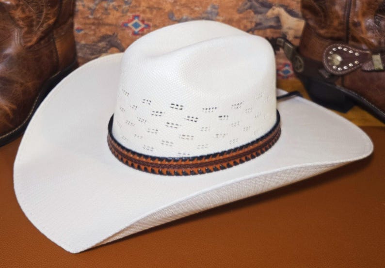 May include: A white cowboy hat with a textured straw-like finish, featuring a ventilated crown and a brown leather hat band with dark stitching. The hat has a wide, curved brim and a classic cattleman crease on the top of the crown.