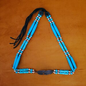 May include: A necklace featuring two strands of bright turquoise cylindrical and cube-shaped beads interspersed with small metallic silver spheres. The strands connect to a central silver-toned metal feather charm and include black leather ties.