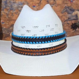May include: A white straw cowboy hat featuring a vented crown and a wide, upturned brim. The hat is adorned with two stacked leather hat bands: one in black with bright blue stitching and one in tan brown with dark brown decorative edge stitching.