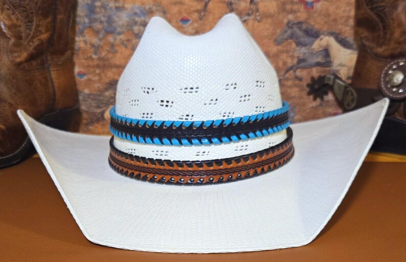 May include: A white straw cowboy hat featuring a vented crown and a wide, upturned brim. The hat is adorned with two stacked leather hat bands: one in black with bright blue stitching and one in tan brown with dark brown decorative edge stitching.