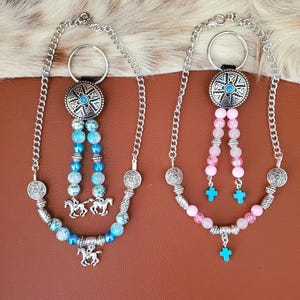 May include: Two beaded keychains with silver-tone metal accents. One features blue and turquoise beads with horse charms, the other pink beads with turquoise crosses. Both have a silver chain and a decorative circular top.