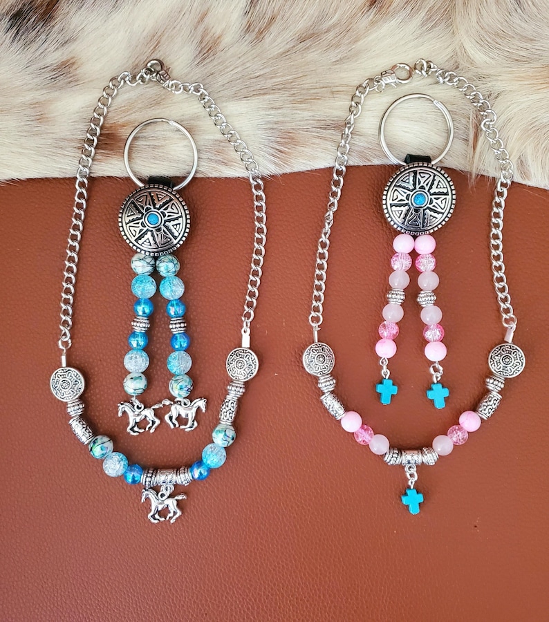 May include: Two beaded keychains with silver-tone metal accents. One features blue and turquoise beads with horse charms, the other pink beads with turquoise crosses. Both have a silver chain and a decorative circular top.