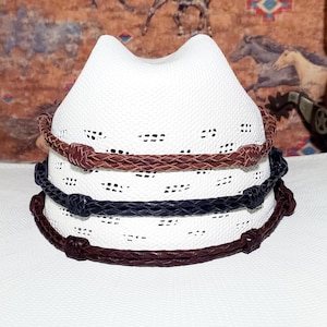 May include: A stack of three white cowboy hats with brown and navy blue braided bands. The hats have a perforated design and a classic cowboy hat shape. The bands are made of a leather-like material.