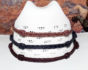 May include: A stack of three white cowboy hats with brown and navy blue braided bands. The hats have a perforated design and a classic cowboy hat shape. The bands are made of a leather-like material.