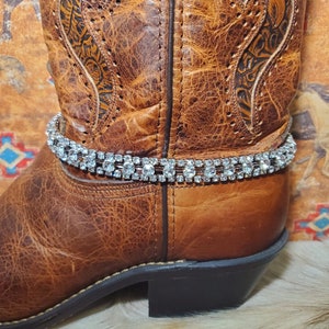 May include: A brown leather cowboy boot with decorative stitching and a silver rhinestone boot chain. The boot has a pointed toe and a stacked heel. The boot chain is made of multiple rows of clear rhinestones.
