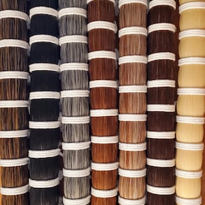 May include: Close-up of eight bundles of horsehair, each tied with white string. The bundles are arranged in a row, and the hair is of different colours, including black, grey, brown, and blonde.