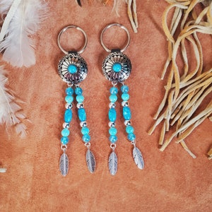 May include: Two silver keychains with turquoise accents. Each keychain features a round, patterned silver pendant with a turquoise center stone, dangling strands of turquoise beads, and silver feather charms. The keychains are displayed on a brown surface.