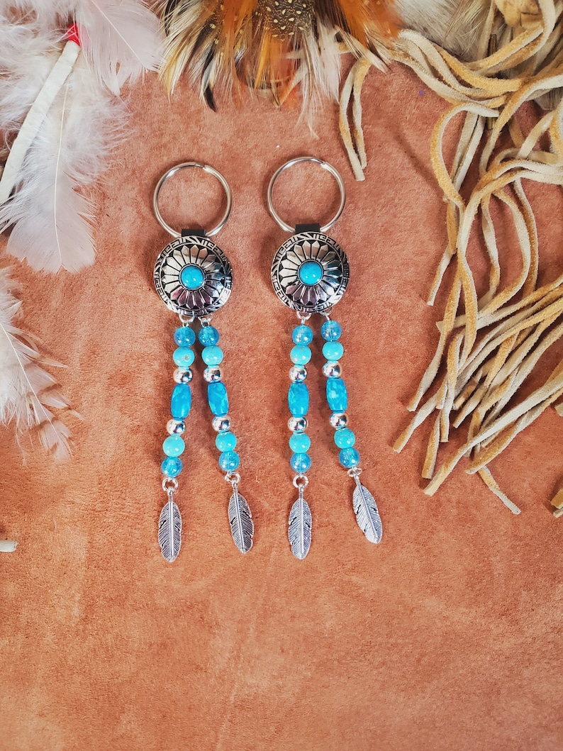 May include: Two silver keychains with turquoise accents. Each keychain features a round, patterned silver pendant with a turquoise center stone, dangling strands of turquoise beads, and silver feather charms. The keychains are displayed on a brown surface.