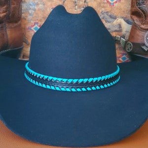 May include: A black felt cowboy hat featuring a wide brim and a high crown. The hat is accented with a decorative band made of black leather interwoven with bright turquoise lacing that wraps around the base of the crown in a repeating pattern.
