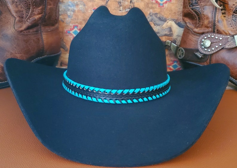 May include: A black felt cowboy hat featuring a wide brim and a high crown. The hat is accented with a decorative band made of black leather interwoven with bright turquoise lacing that wraps around the base of the crown in a repeating pattern.