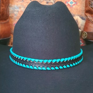May include: A black felt hat featuring a decorative hatband. The band consists of a central black leather strip with an embossed braided pattern, flanked by two rows of bright turquoise leather lacing woven through the edges of the band.