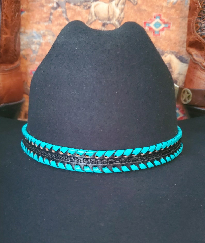 May include: A black felt hat featuring a decorative hatband. The band consists of a central black leather strip with an embossed braided pattern, flanked by two rows of bright turquoise leather lacing woven through the edges of the band.