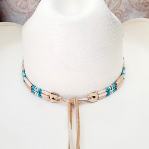 May include: A white cowboy hat with a brown leather hat band decorated with turquoise and silver beads. The hat band has a long brown leather tassel.