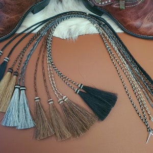 May include: A set of eight braided leather boot laces with tassels in various colors, including black, brown, gray, and white. The laces are arranged in a semi-circle on a brown surface.