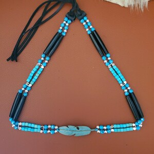 May include: A turquoise and black beaded necklace with a turquoise feather pendant. The necklace is made with small, square turquoise beads and black beads. The feather pendant is a light turquoise colour and has a silver accent.