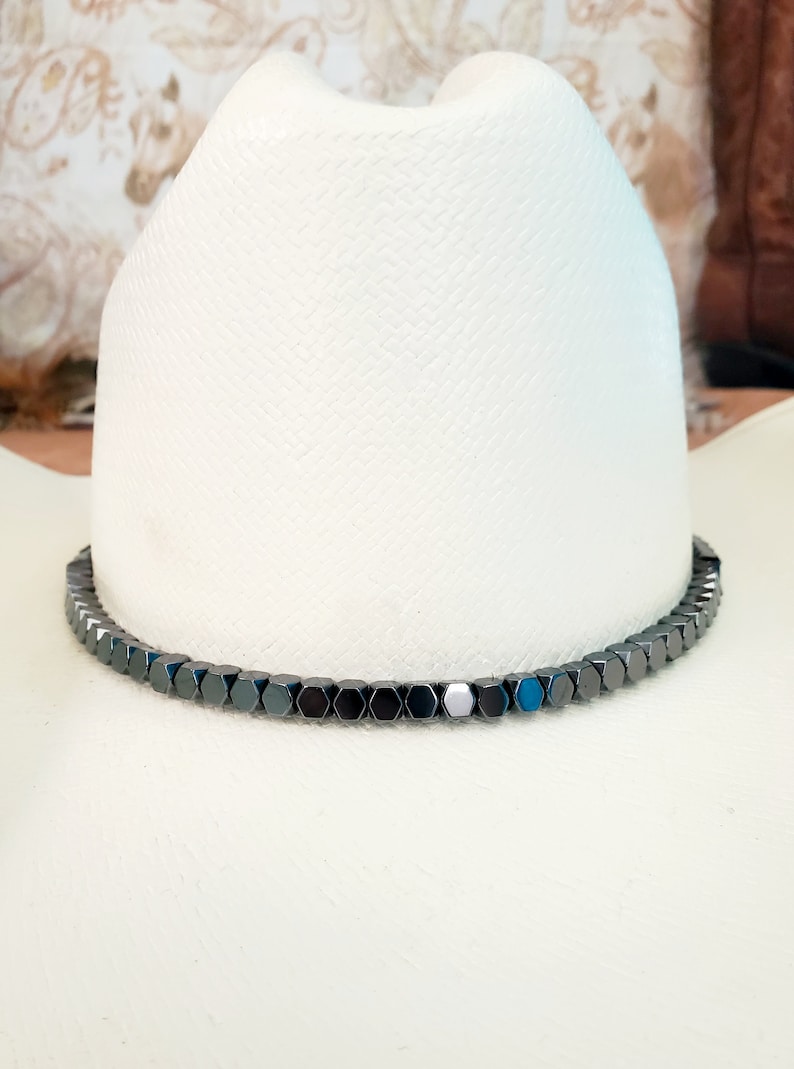 Pu&ograve; includere: Un cappello da cowboy bianco con una fascia di perline esagonali nere attorno alla base della corona.