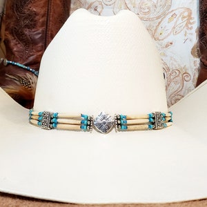May include: A white cowboy hat with a turquoise and silver hat band. The hat band has a silver heart-shaped charm in the center.
