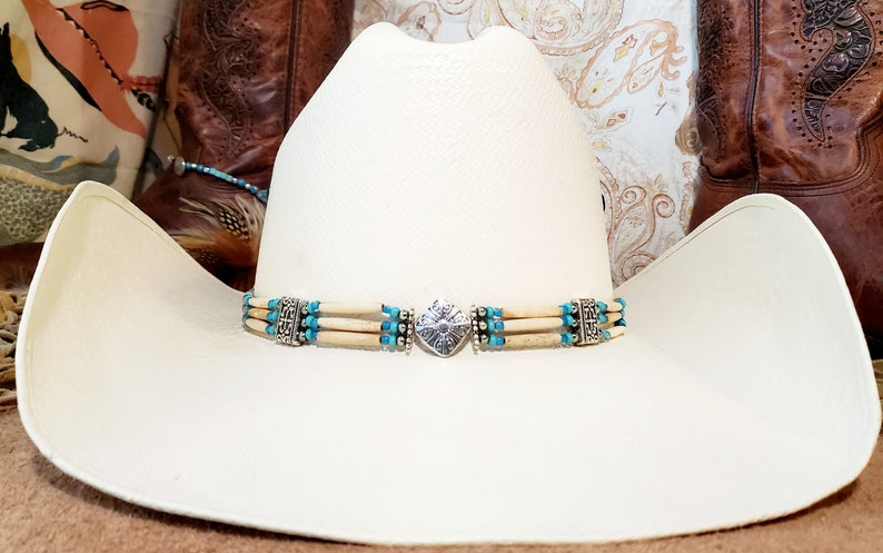May include: A white cowboy hat with a turquoise and silver hat band. The hat band has a silver heart-shaped charm in the center.