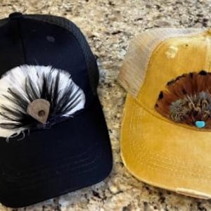 Puede incluir: Dos gorras de camionero, una negra y otra amarillo mostaza, cada una adornada con un dise&ntilde;o de plumas decorativas. La gorra negra presenta un abanico de plumas blancas y negras con un acento de cuero marr&oacute;n. La gorra amarilla tiene un dise&ntilde;o de plumas marrones y beige con un acento turquesa.