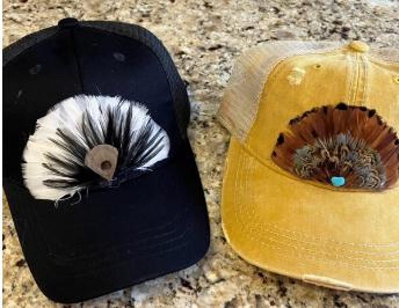 Puede incluir: Dos gorras de camionero, una negra y otra amarillo mostaza, cada una adornada con un dise&ntilde;o de plumas decorativas. La gorra negra presenta un abanico de plumas blancas y negras con un acento de cuero marr&oacute;n. La gorra amarilla tiene un dise&ntilde;o de plumas marrones y beige con un acento turquesa.