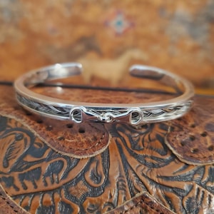 May include: A silver bangle bracelet with a braided horsehair band and a silver horse bit charm. The bracelet is on a brown leather background.