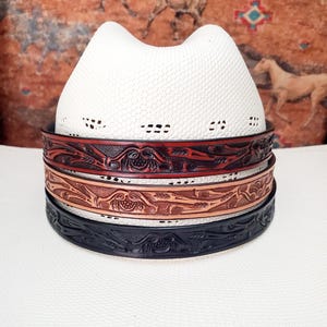 Può includere: Un cappello da cowboy bianco con una trama intrecciata, che mostra tre fasce decorative in pelle. Le fasce sono nei toni del rosso, marrone e nero, ognuna con un design floreale in rilievo. Il cappello e le fasce sono su uno sfondo a motivi.