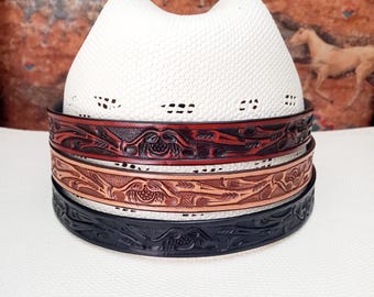May include: A white cowboy hat with a woven texture, showcasing three decorative leather hat bands. The bands are in shades of red, tan, and black, each featuring an embossed floral design. The hat and bands are set against a patterned background.