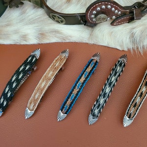May include: Five hair clips with different patterns and colors. The clips are made of leather and have a silver metal accent. The patterns include black and white, brown and white, and blue and white.