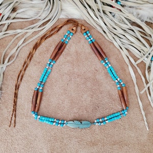 May include: A turquoise and brown beaded necklace with a silver feather charm. The necklace is on a light brown suede background.