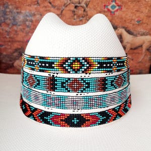 May include: A white hat with a decorative band featuring a colourful beaded design. The band showcases patterns in turquoise, red, black, yellow, and white. The hat has a unique, curved crown.