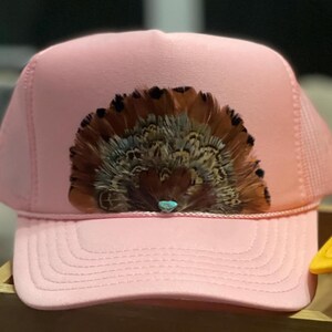 Puede incluir: Gorra de camionero rosa con un arreglo decorativo de plumas marrones y beige, acentuado con una peque&ntilde;a piedra turquesa. La gorra tiene una parte trasera de malla y una visera curva.