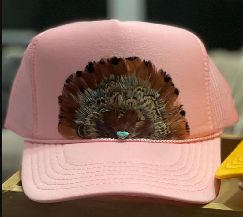 Puede incluir: Gorra de camionero rosa con un arreglo decorativo de plumas marrones y beige, acentuado con una peque&ntilde;a piedra turquesa. La gorra tiene una parte trasera de malla y una visera curva.