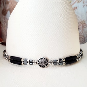 May include: A white cowboy hat with a black and silver beaded hat band. The hat band has a silver sunburst design in the centre.
