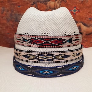Op de afbeelding: Een witte geweven hoed met drie decoratieve banden. De banden hebben geometrische patronen in rood, zwart, blauw en beige. De hoed heeft een klassieke fedora-vorm.