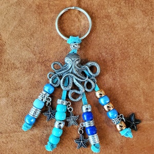 Peut inclure: Un porte-clés en forme de pieuvre argenté avec des perles bleues et dorées et des breloques en forme d'étoile.