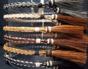May include: A collection of six braided horsehair hatbands, each featuring a distinct color pattern of black, white, and various shades of brown. Every band is finished with a decorative knot and a long, flowing horsehair tassel at the end.