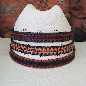 May include: A white straw cowboy hat featuring a cattleman crease and a decorative hat band. The band consists of three stacked leather layers in alternating shades of dark brown and tan, each featuring intricate braided and serrated edge details.