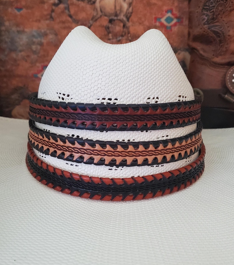 May include: A white straw cowboy hat featuring a cattleman crease and a decorative hat band. The band consists of three stacked leather layers in alternating shades of dark brown and tan, each featuring intricate braided and serrated edge details.