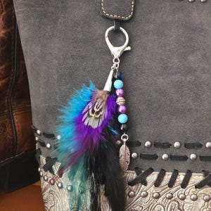 May include: A silver charm with a black feather and turquoise and purple beads attached to a silver clasp. The charm is hanging from a gray bag with silver studs.