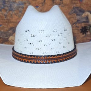 May include: A white cowboy hat featuring a ventilated crown with a textured weave pattern. The hat includes a decorative band made of brown leather with black stitched edges and a braided center detail, set against a wide, curved brim.