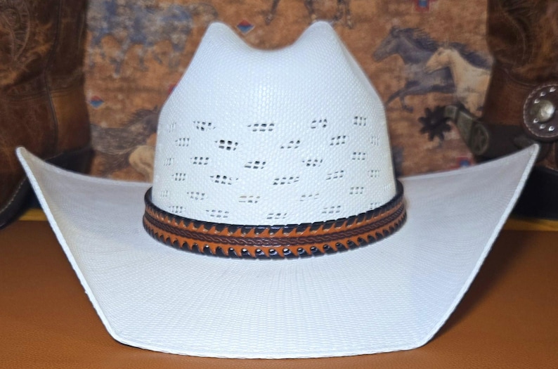 May include: A white cowboy hat featuring a ventilated crown with a textured weave pattern. The hat includes a decorative band made of brown leather with black stitched edges and a braided center detail, set against a wide, curved brim.