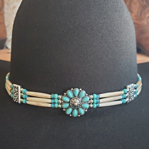 May include: Black cowboy hat with a decorative hat band. The hat band features three strands of off-white beads, turquoise accents, and a silver-toned floral centerpiece. The hat band is secured with silver-toned clasps.