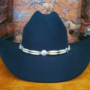 May include: A black cowboy hat with a white bone and silver bead trim. The hat has a wide brim and a tall crown.