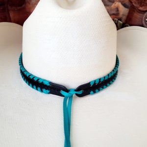 May include: A white cowboy hat featuring a decorative hat band made of black leather braided with bright turquoise cord. The band includes a central knot detail with two long turquoise leather ties hanging down from the front of the hat crown.