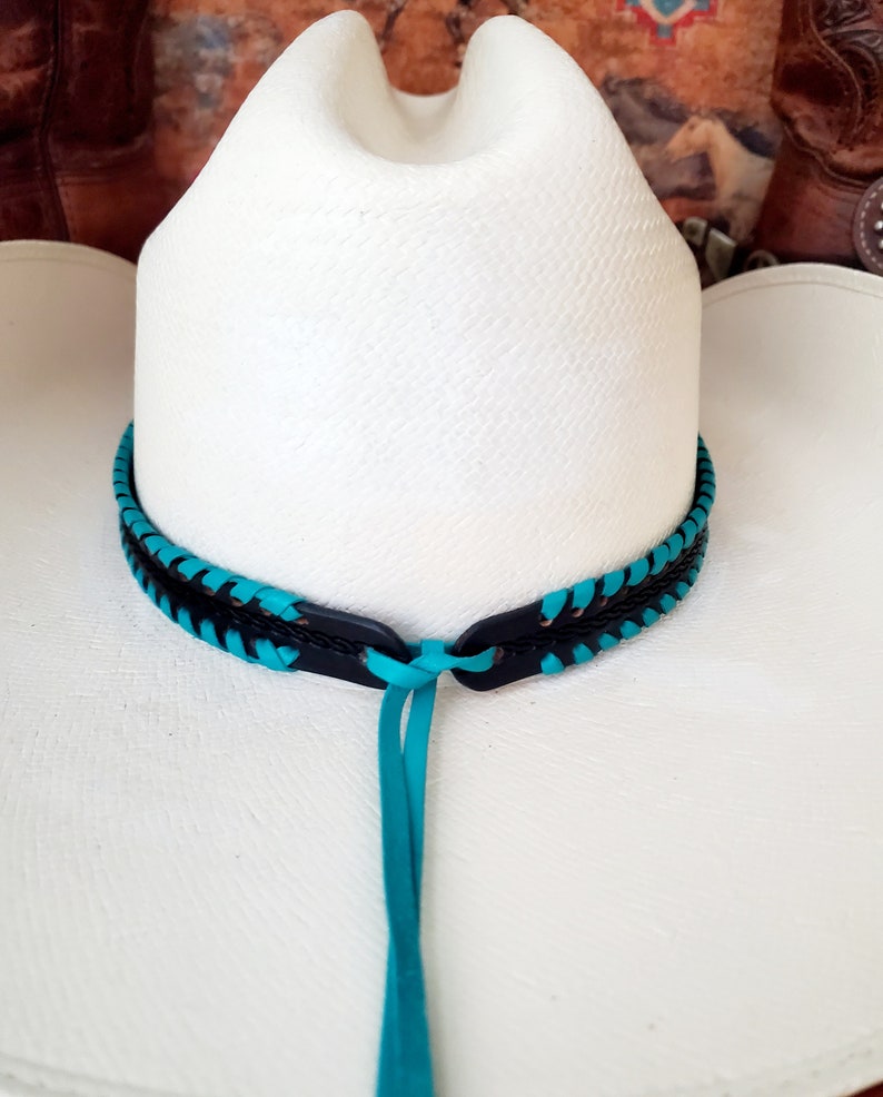 May include: A white cowboy hat featuring a decorative hat band made of black leather braided with bright turquoise cord. The band includes a central knot detail with two long turquoise leather ties hanging down from the front of the hat crown.