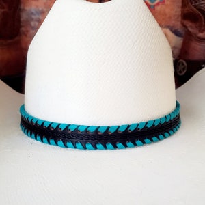May include: A white cowboy hat featuring a textured straw-like finish and a cattleman crease. The hat is accented with a decorative hatband composed of black leather braided with vibrant turquoise leather lacing around the base of the crown.