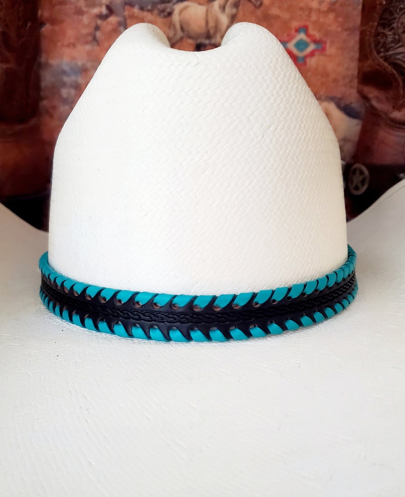 May include: A white cowboy hat featuring a textured straw-like finish and a cattleman crease. The hat is accented with a decorative hatband composed of black leather braided with vibrant turquoise leather lacing around the base of the crown.