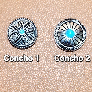 May include: Two silver-toned conchos with turquoise-colored centers. The conchos are round with detailed geometric designs. The text "Concho 1" and "Concho 2" are below each concho.