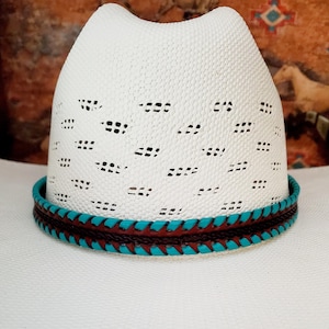 May include: A white cowboy hat featuring a ventilated crown with a repeating pattern of small rectangular cutouts. The base of the crown is encircled by a decorative hat band composed of braided dark brown leather and bright turquoise leather.